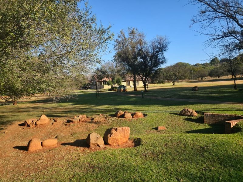 A spacious, well-maintained residential garden featuring mature trees, natural rock landscaping, and a brick retaining wall on a large vacant land plot.
