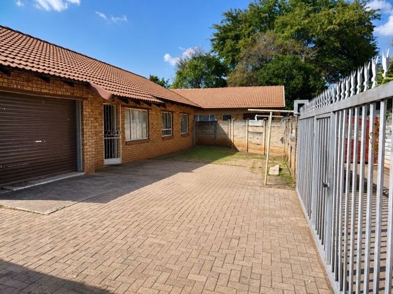 Front exterior view of a brick house featuring a paved driveway, metal garage door, and secure perimeter fencing.