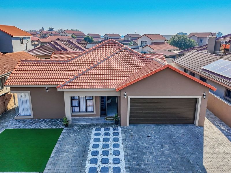 Aerial view of a well-maintained residential property featuring a tiled roof, paved driveway, integrated garage, and a neat artificial lawn area.