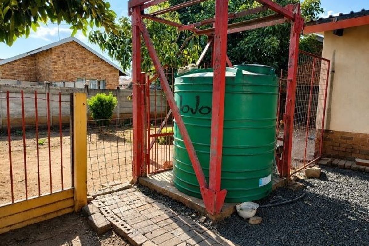 Rear exterior area featuring a large green water storage tank mounted on a red metal support frame, situated on a paved section of the yard.
