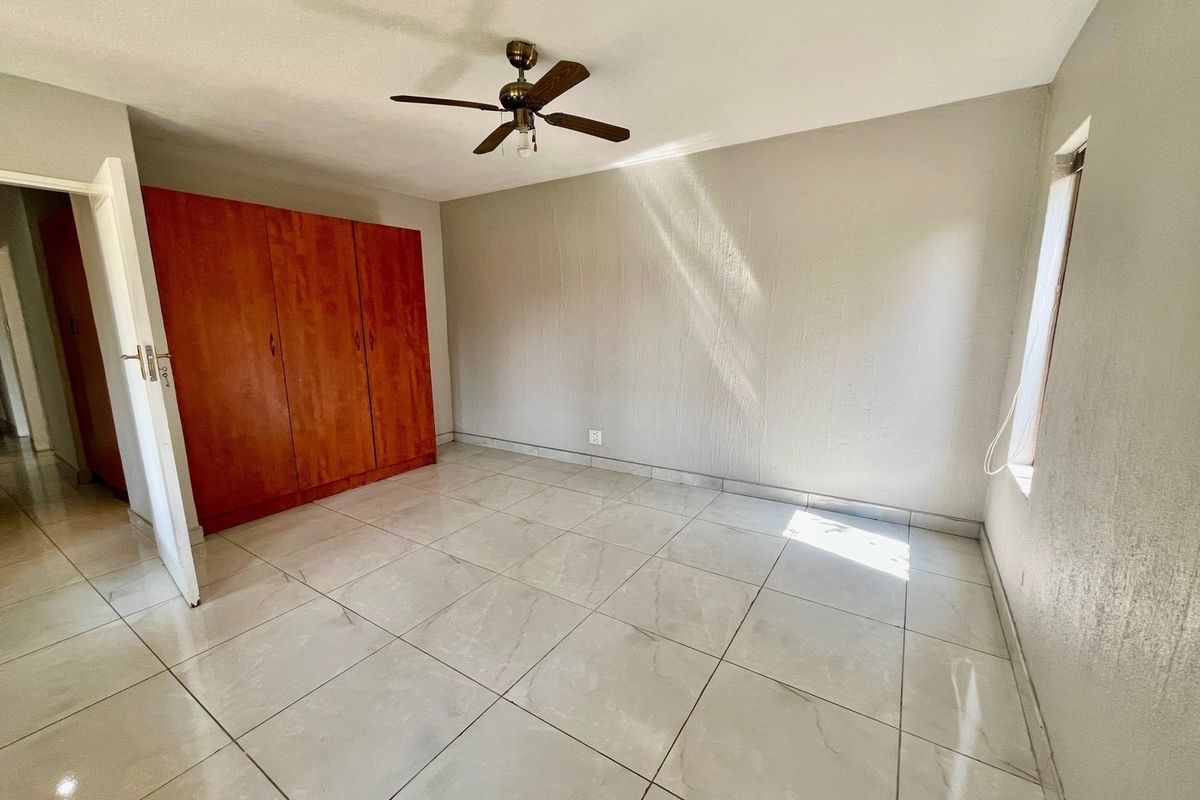 A bright, empty bedroom featuring neutral tiled flooring, built-in wooden cupboards, and a ceiling fan.
