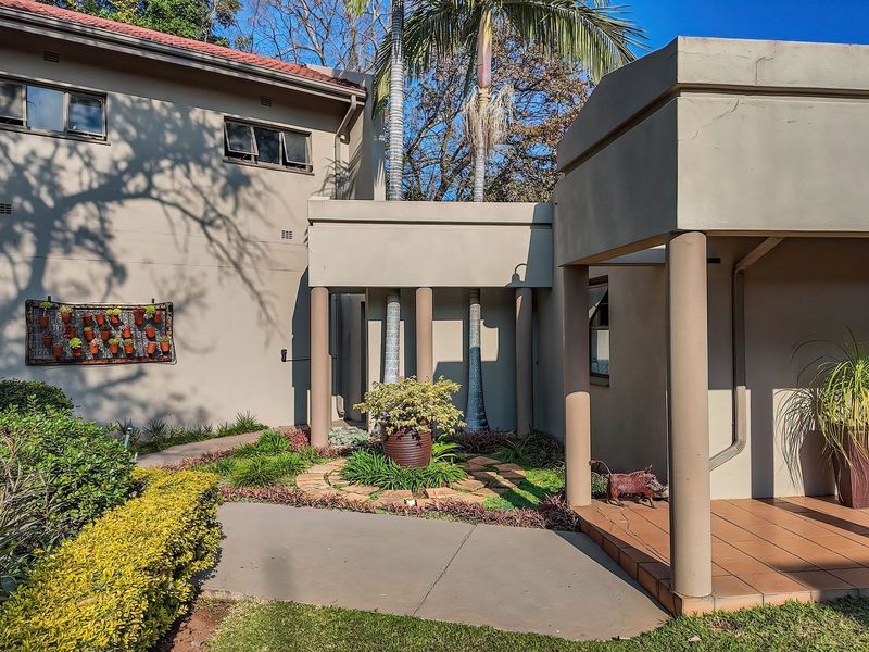 Front entrance area featuring a covered tiled patio, concrete walkway, and established garden landscaping with mature trees.
