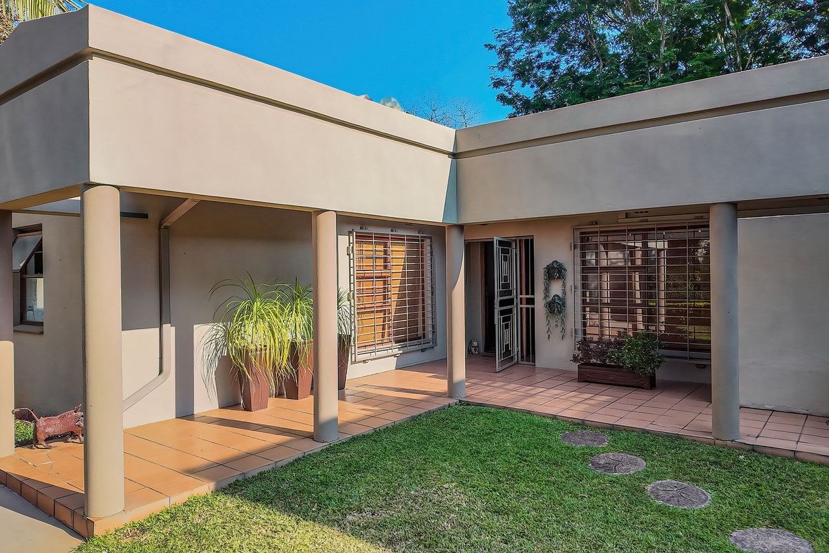 A covered patio area featuring tiled flooring, concrete support columns, and security bars on the windows, overlooking a small garden space.