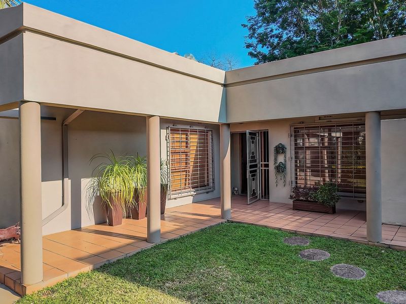 A covered patio area featuring tiled flooring, concrete support columns, and security bars on the windows, overlooking a small garden space.