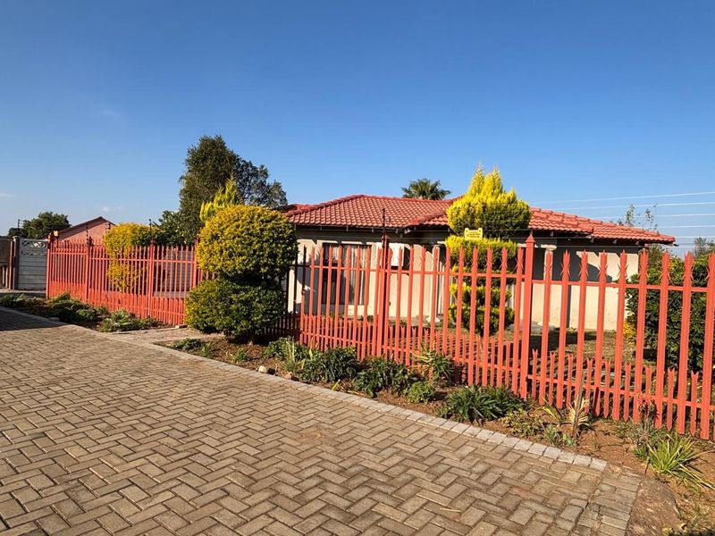 Front view of the property featuring a paved driveway, established garden beds, and secure red metal palisade fencing.