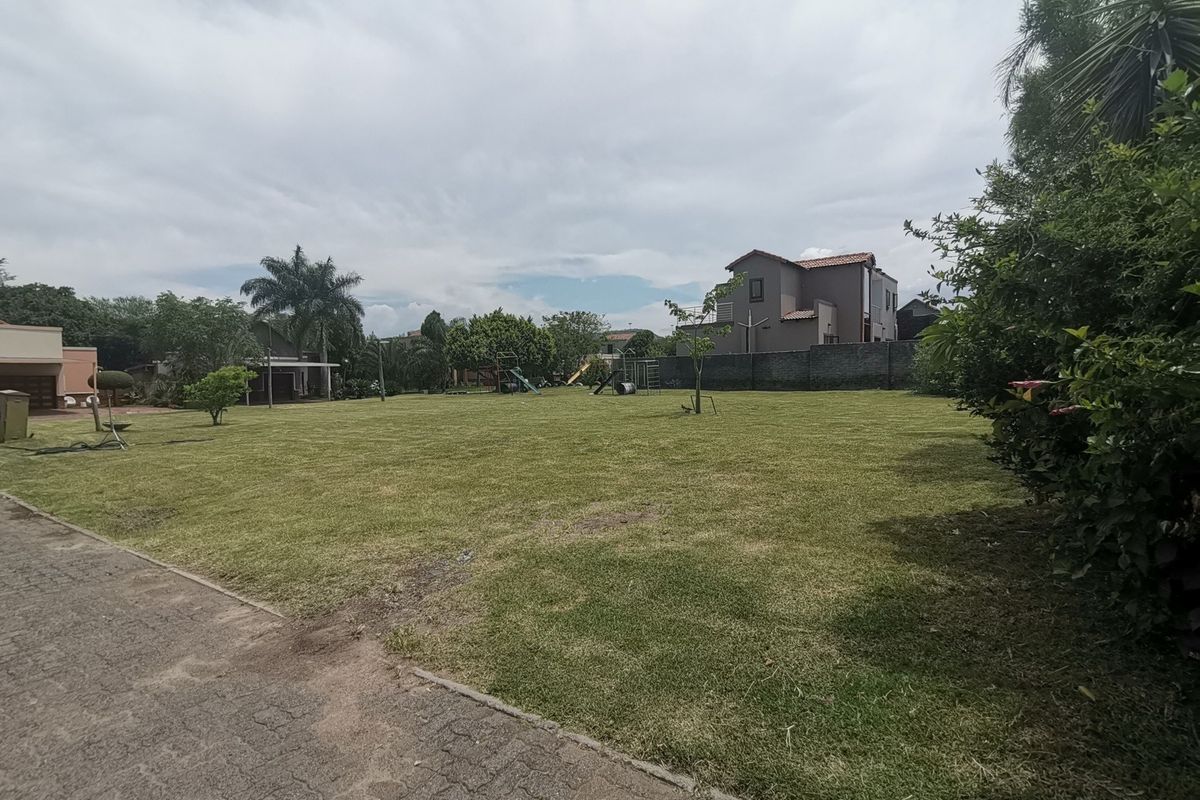 A spacious, well-maintained residential garden featuring a large lawn area, paved driveway access, and perimeter boundary walls.