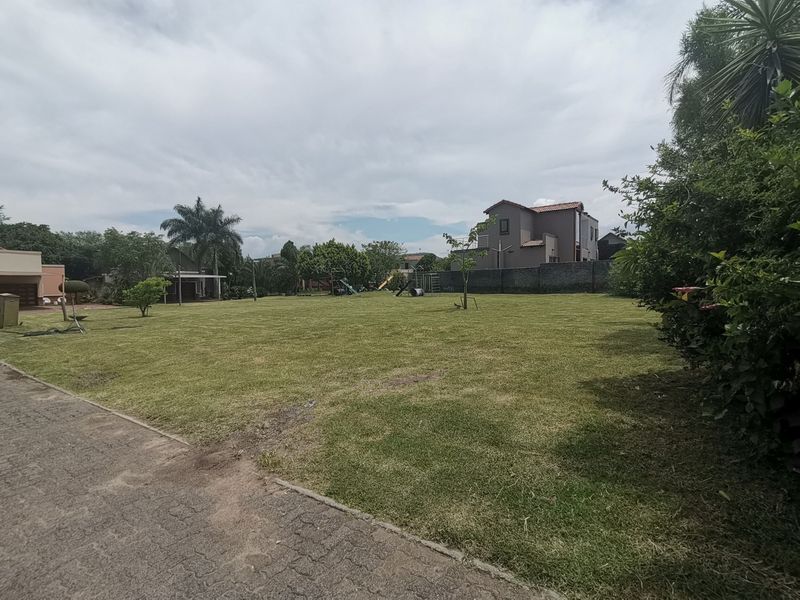 A spacious, well-maintained residential garden featuring a large lawn area, paved driveway access, and perimeter boundary walls.