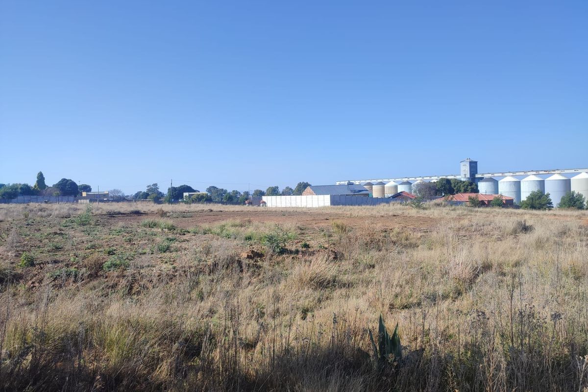 A view of a vacant residential plot featuring dry grass and a perimeter wall, with industrial silos visible in the background.