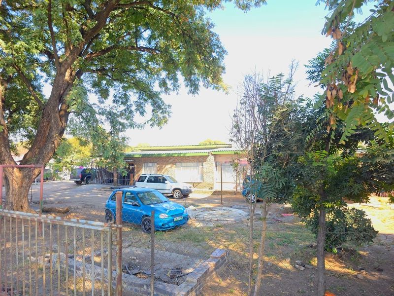 Front view of a single-story brick house featuring a metal roof, set behind a wire mesh fence with mature trees and a large yard.
