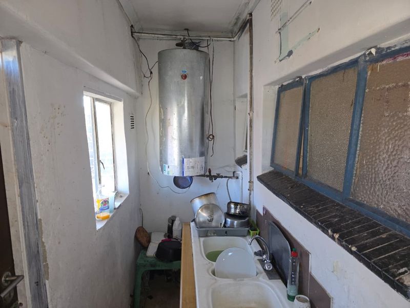 A functional scullery area featuring a double basin sink, wall-mounted electric water heater, and basic utility finishes in need of maintenance and cosmetic updates.
