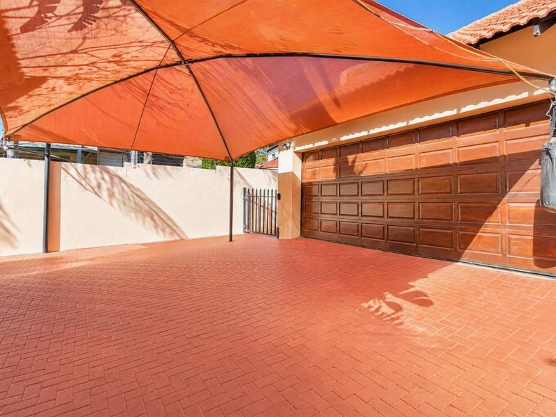 A paved driveway area featuring a large orange shade sail and a wooden sectional garage door, enclosed by a perimeter wall.