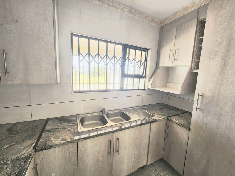 A functional kitchen featuring light wood-grain cabinetry, dark marble-effect laminate countertops, a double stainless steel sink, and security bars on the window.