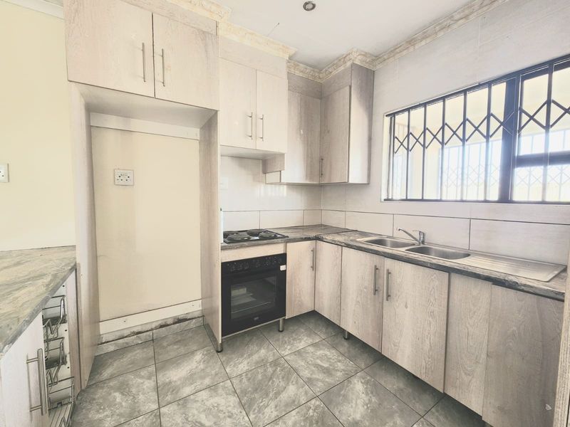 A functional kitchen featuring light wood-grain cabinetry, laminate countertops, a built-in oven and hob, and tiled flooring with security bars on the window.