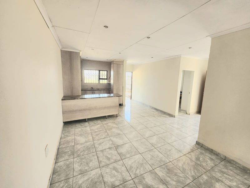 An open-plan living and kitchen area featuring neutral tiled flooring, modern light-toned cabinetry, and a functional kitchen island in a clean, vacant space.