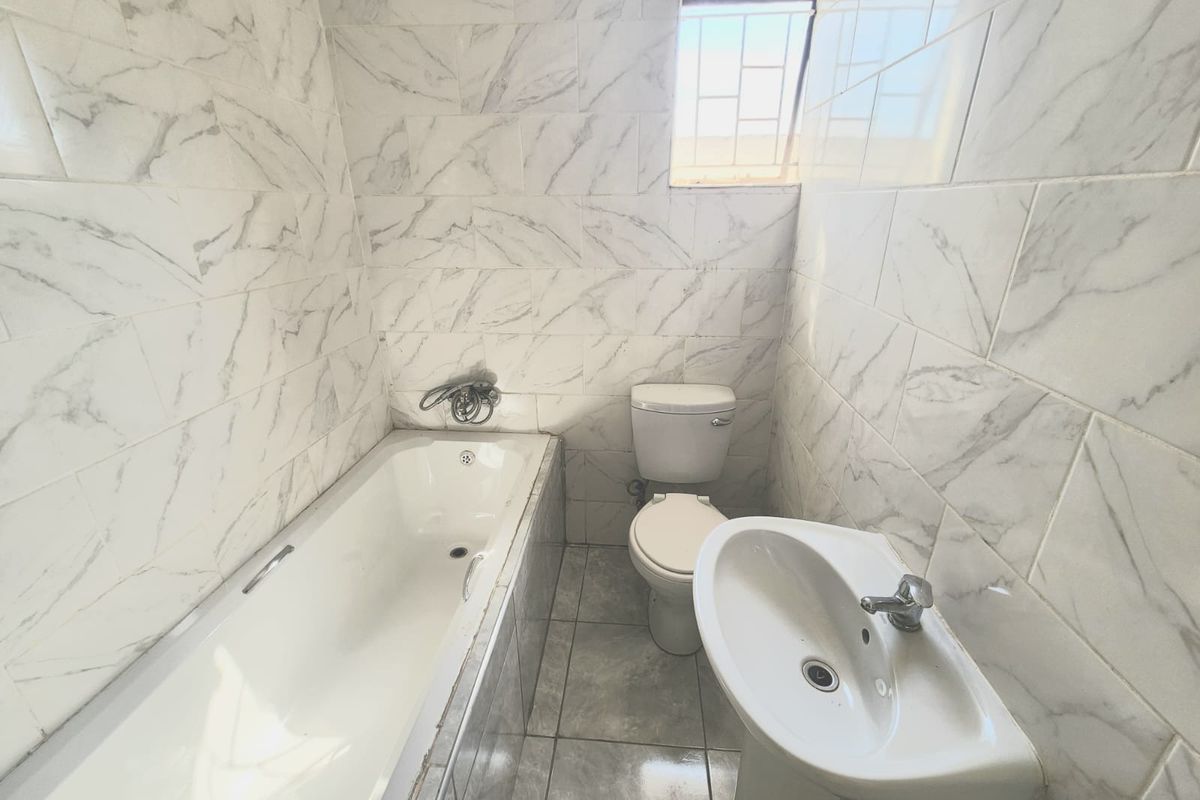 A compact bathroom featuring marble-look wall tiles, a standard bathtub, pedestal sink, and toilet with a security-grille window.