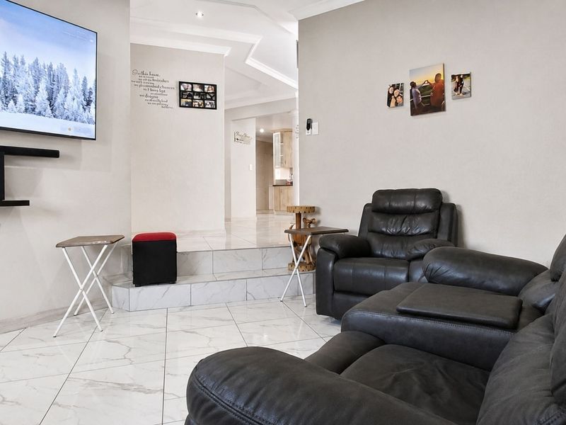 A modern living room featuring polished marble-look floor tiles, a raised platform area, and recessed ceiling lighting, leading through to an open-plan kitchen space.