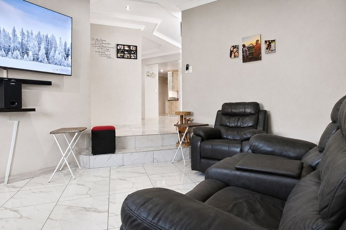 A modern living room featuring polished marble-look floor tiles, a raised platform area, and recessed ceiling lighting, leading through to an open-plan kitchen space.