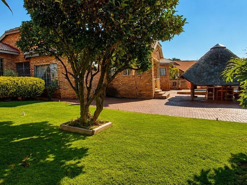 A spacious rear garden featuring a well-maintained lawn, brick-paved patio area, and a traditional thatched lapa, ideal for outdoor entertaining.