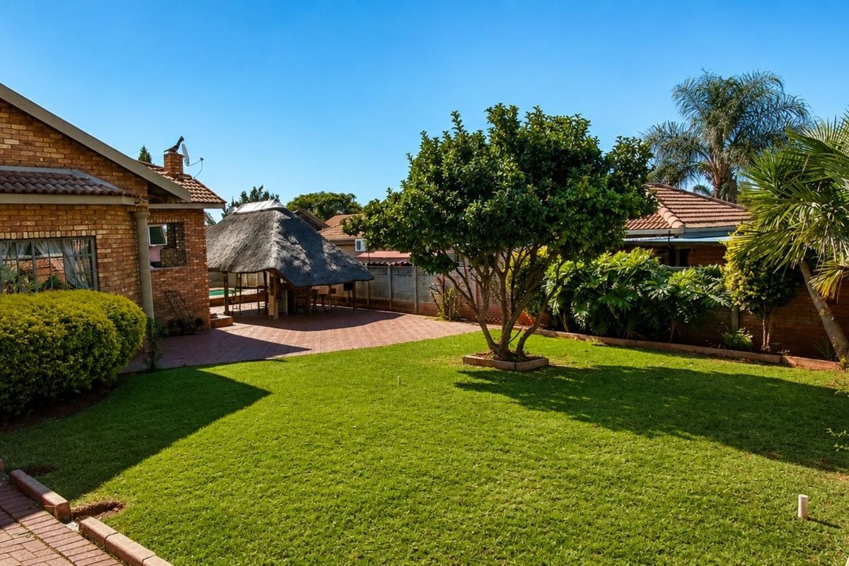 A well-maintained rear garden featuring a brick house, a paved patio area, and a traditional thatched lapa structure.