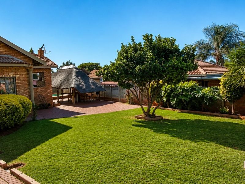 A well-maintained rear garden featuring a brick house, a paved patio area, and a traditional thatched lapa structure.
