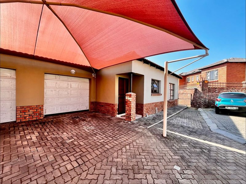 Front exterior view of the townhouse featuring a paved driveway, garage access, and a large red shade sail covering the parking area.