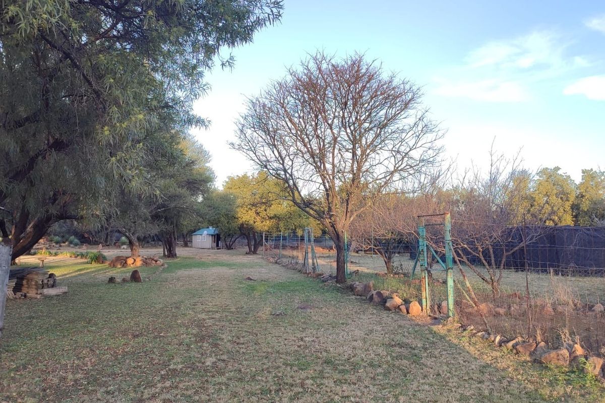 A spacious farm garden area featuring mature trees, wire fencing, and natural stone borders, offering a rustic outdoor setting.