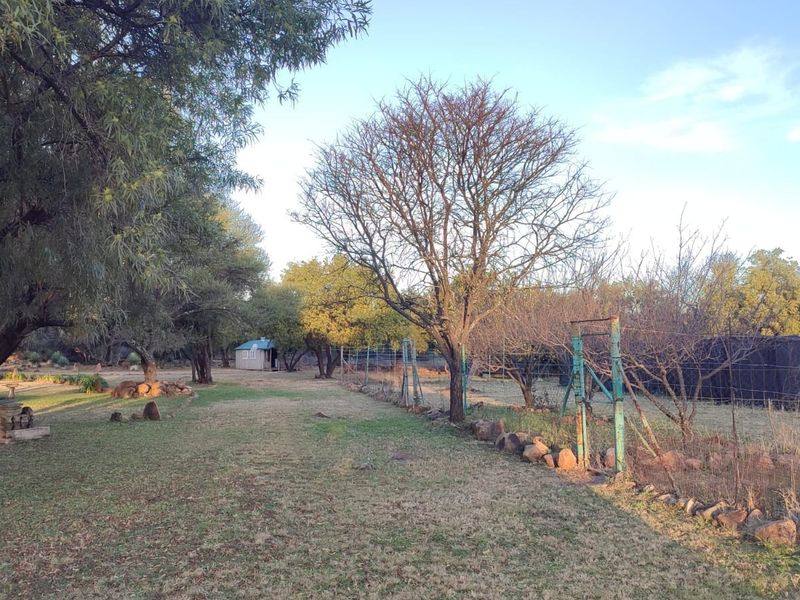 A spacious farm garden area featuring mature trees, wire fencing, and natural stone borders, offering a rustic outdoor setting.