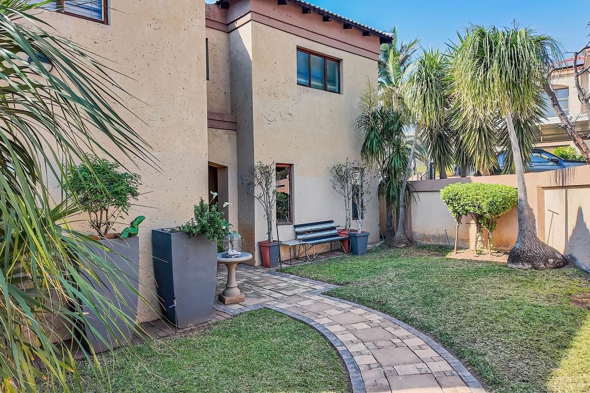A private rear garden featuring a paved walkway, established landscaping, and a stucco townhouse facade with a boundary wall.