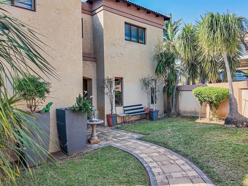 A private rear garden featuring a paved walkway, established landscaping, and a stucco townhouse facade with a boundary wall.