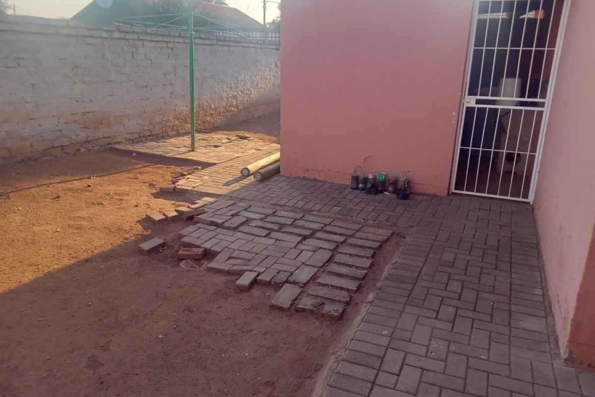 Rear exterior area featuring a paved walkway, security gate, and perimeter wall, with some uneven paving and exposed ground requiring maintenance.
