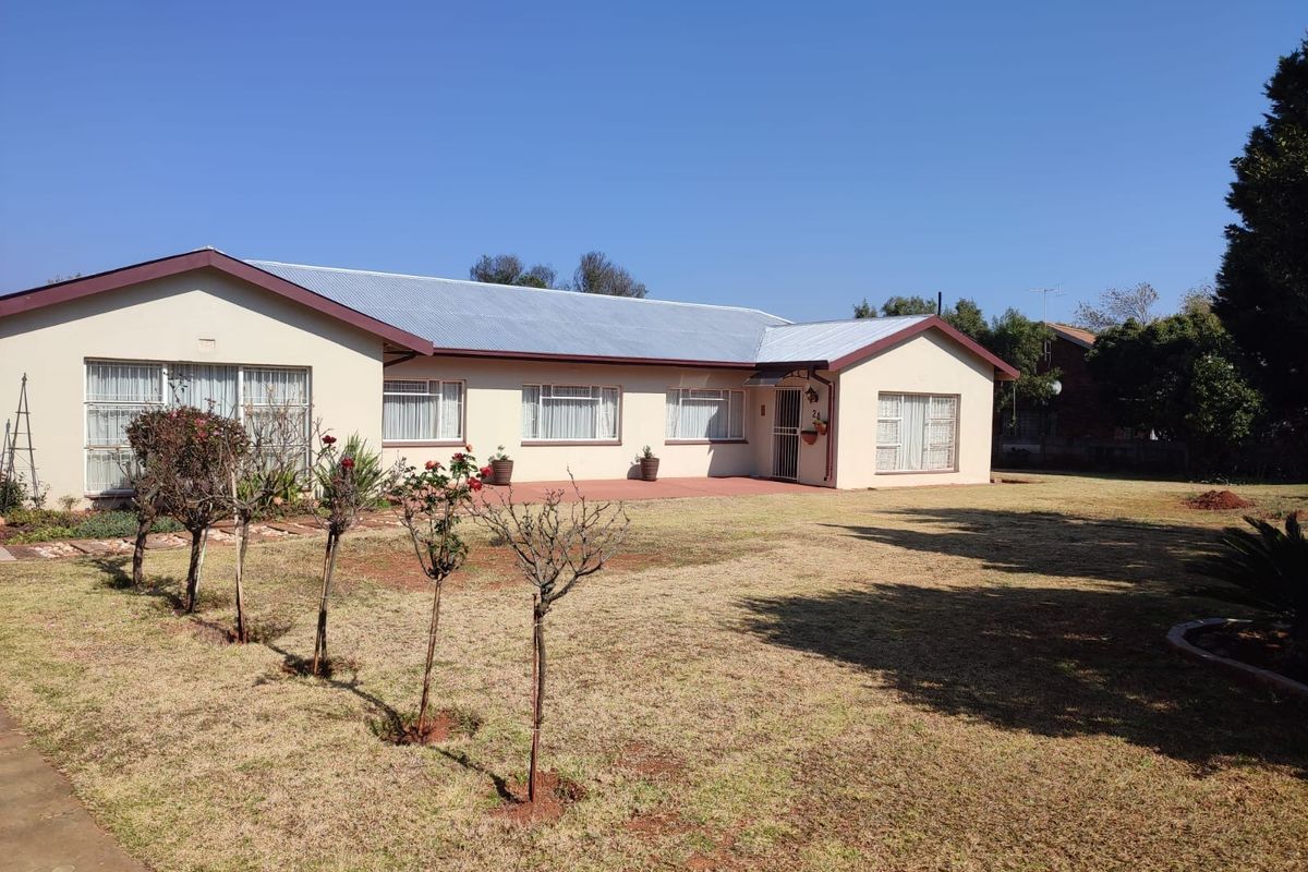 A single-story residential property featuring a corrugated metal roof, paved patio area, and a spacious lawn with established garden beds.