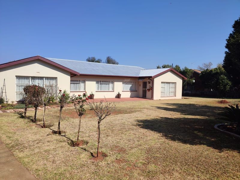 A single-story residential property featuring a corrugated metal roof, paved patio area, and a spacious lawn with established garden beds.