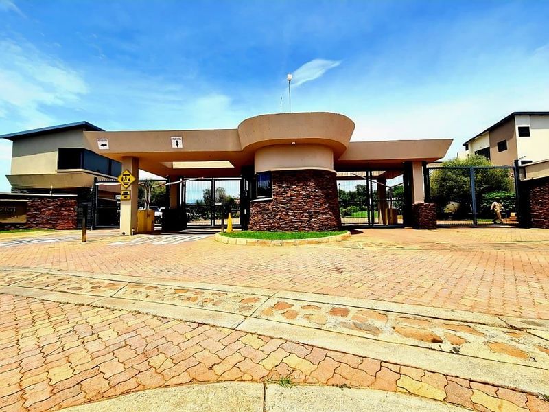 A well-maintained residential estate entrance featuring a modern security gatehouse, automated boom gates, and stone-clad architectural elements.