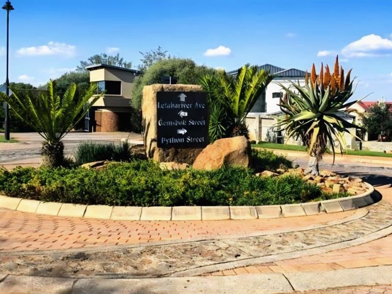 A well-maintained residential street roundabout featuring landscaped gardens, stone signage, and paved surfaces within a secure estate environment.