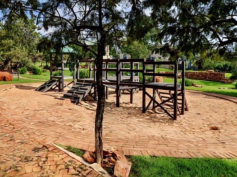 A well-maintained residential garden featuring a wooden playground structure set within a sand pit, surrounded by brick paving and established lawn.