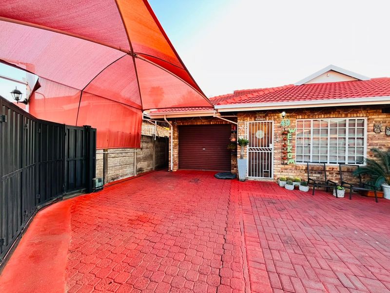 Front exterior view featuring a brick facade, tiled roof, paved driveway, and a large red shade sail covering the parking area.
