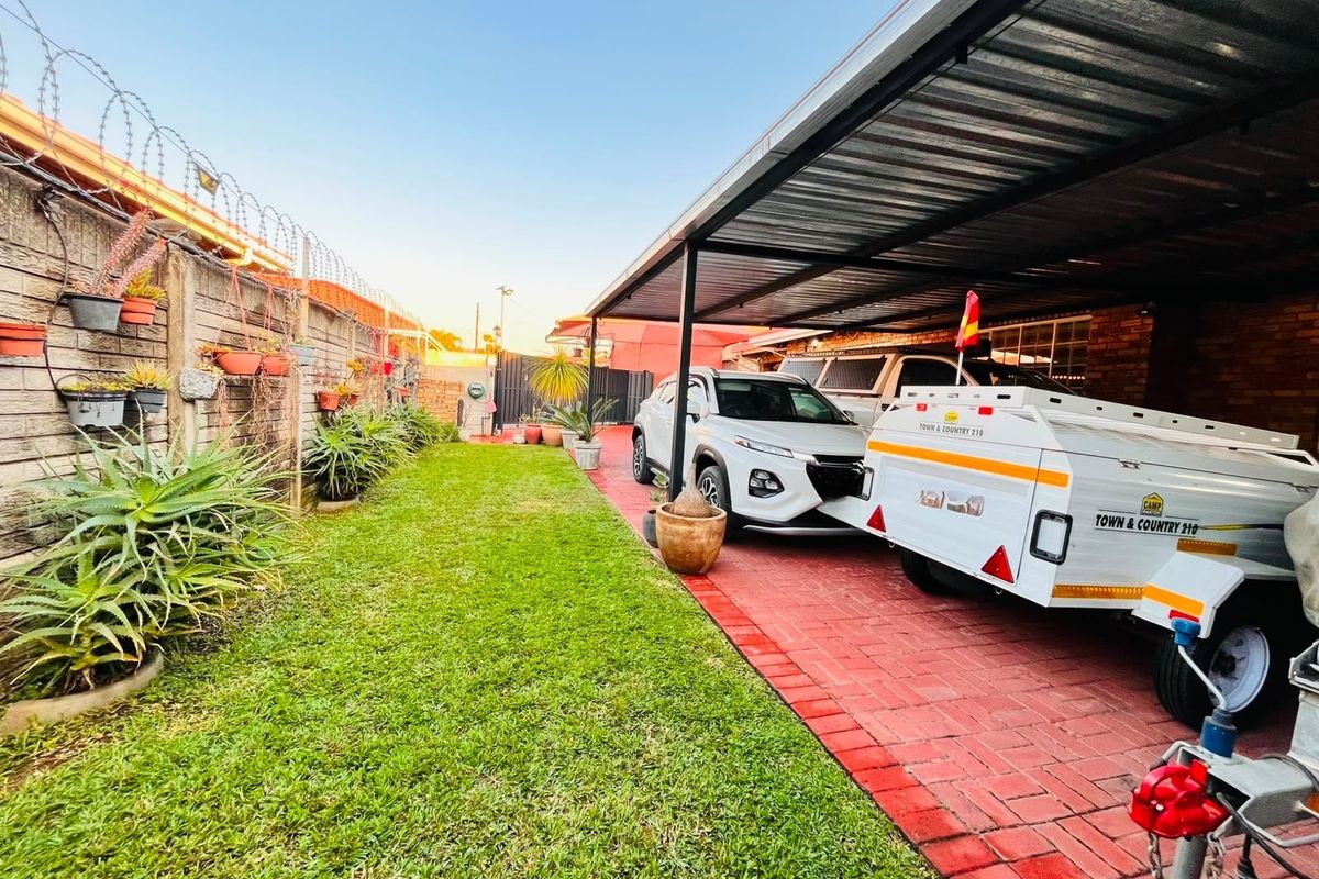 A rear exterior view featuring a paved driveway, a covered carport structure, and a boundary wall with security fencing.