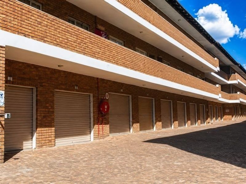 Exterior view of a multi-unit apartment complex featuring a row of individual garages with roller shutter doors and paved driveway access.