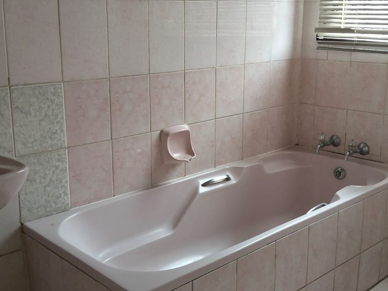 A dated bathroom featuring a pink built-in bathtub and matching pedestal basin with ceramic wall tiling and a window with venetian blinds.