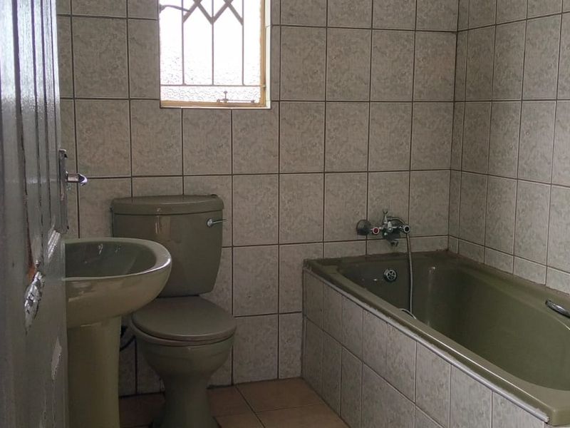 A functional bathroom featuring a pedestal basin, toilet, and bathtub with full-height wall tiling and a window with security bars.