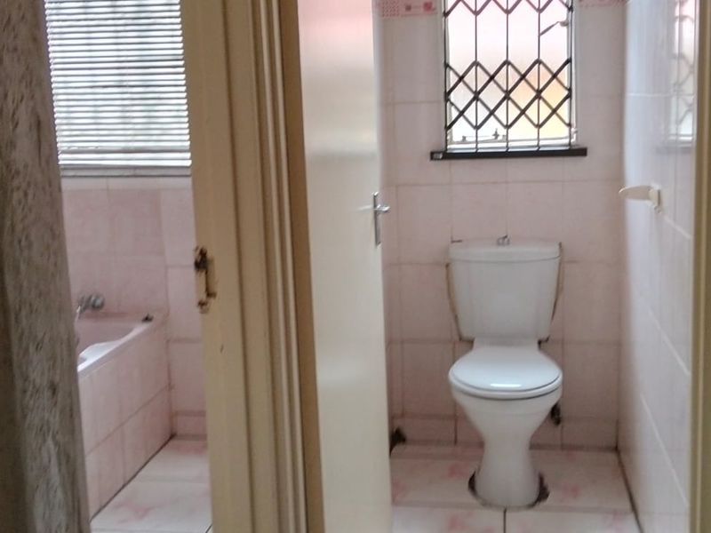 A dated bathroom featuring a separate toilet and bathtub area with tiled finishes and security bars on the window.