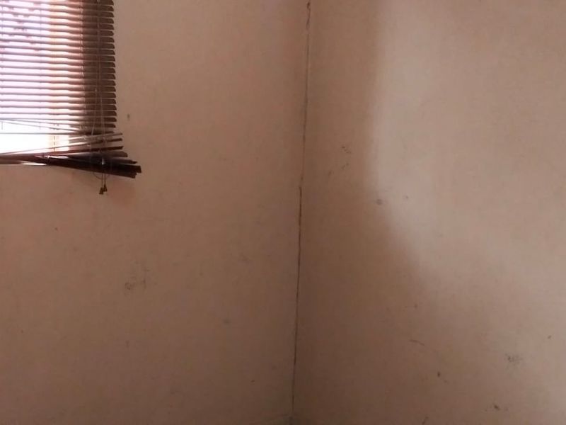 A corner view of an empty room featuring a window with damaged venetian blinds and visible wall cracks requiring maintenance.