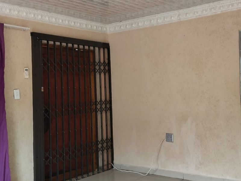 Open plan living area featuring tiled floors, decorative ceiling cornices, and a security gate, with an adjacent kitchen visible through the doorway.