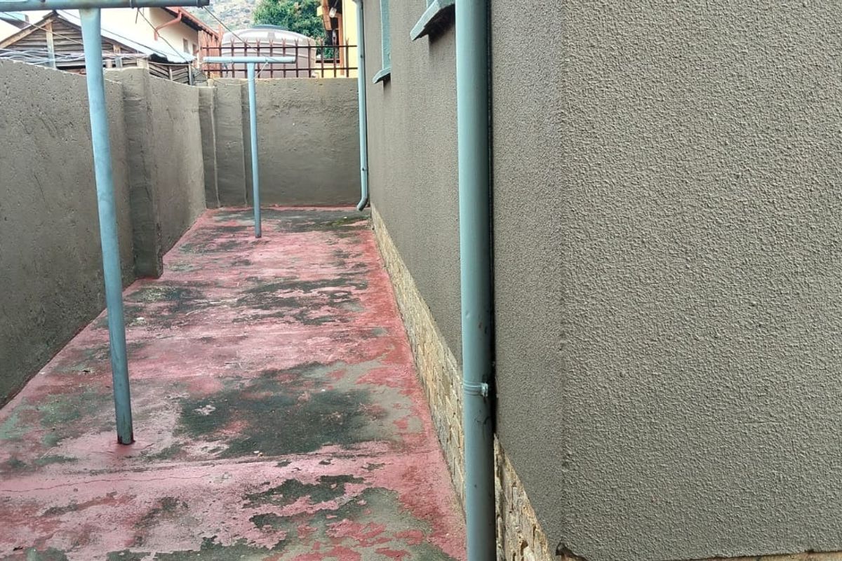 A side exterior area featuring a boundary wall, downpipe, and concrete paving with visible surface wear and peeling paint.