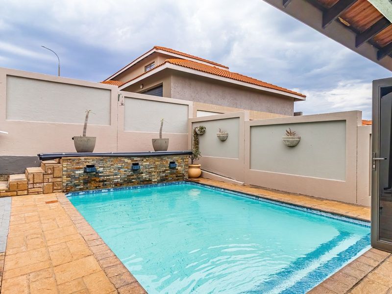 A well-maintained outdoor pool area featuring a stone-clad water feature, paved decking, and a secure perimeter wall, adjacent to a covered patio space.