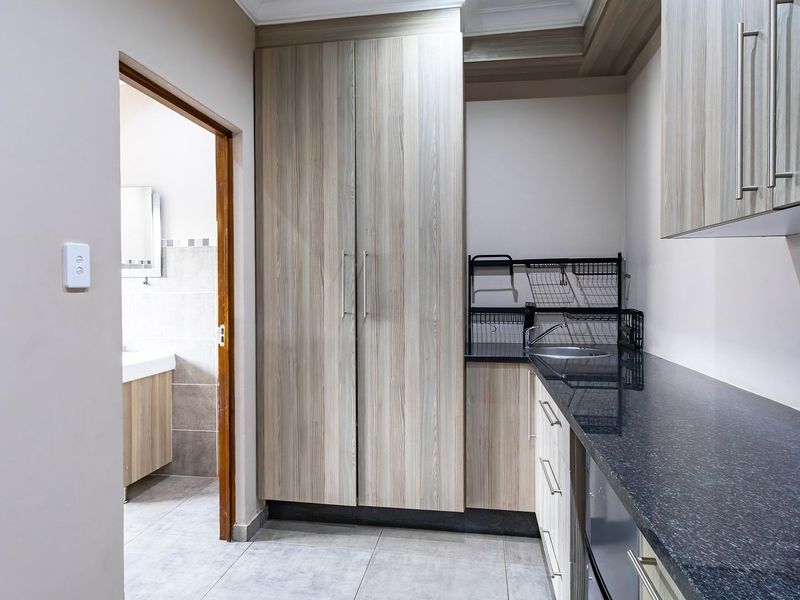 A clean and functional scullery featuring modern wood-grain cabinetry, granite countertops, a stainless steel sink, and neutral tiled flooring with direct access to a bathroom.