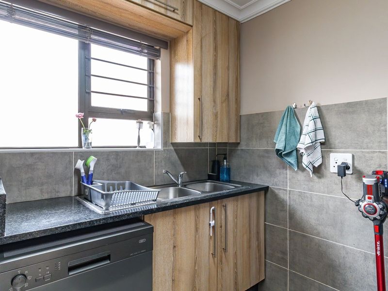 A functional scullery featuring wood-look cabinetry, granite countertops, a double sink, and modern grey tiled splashbacks.
