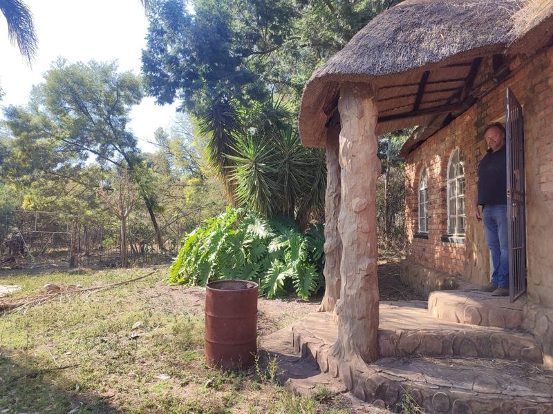Exterior view of a rustic farm building featuring a traditional thatch roof, stone-clad pillars, and arched windows set within a natural garden landscape.