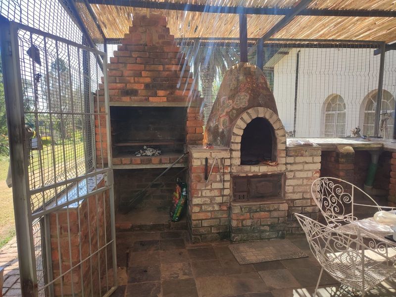 Covered outdoor entertainment area featuring a traditional built-in brick braai and a pizza oven, set under a rustic thatched roof.