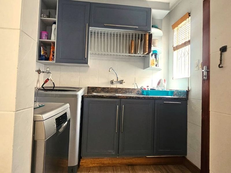 A functional laundry area featuring dark grey cabinetry, a stone-look countertop, stainless steel sink, and tiled splashback with dedicated space for appliances.